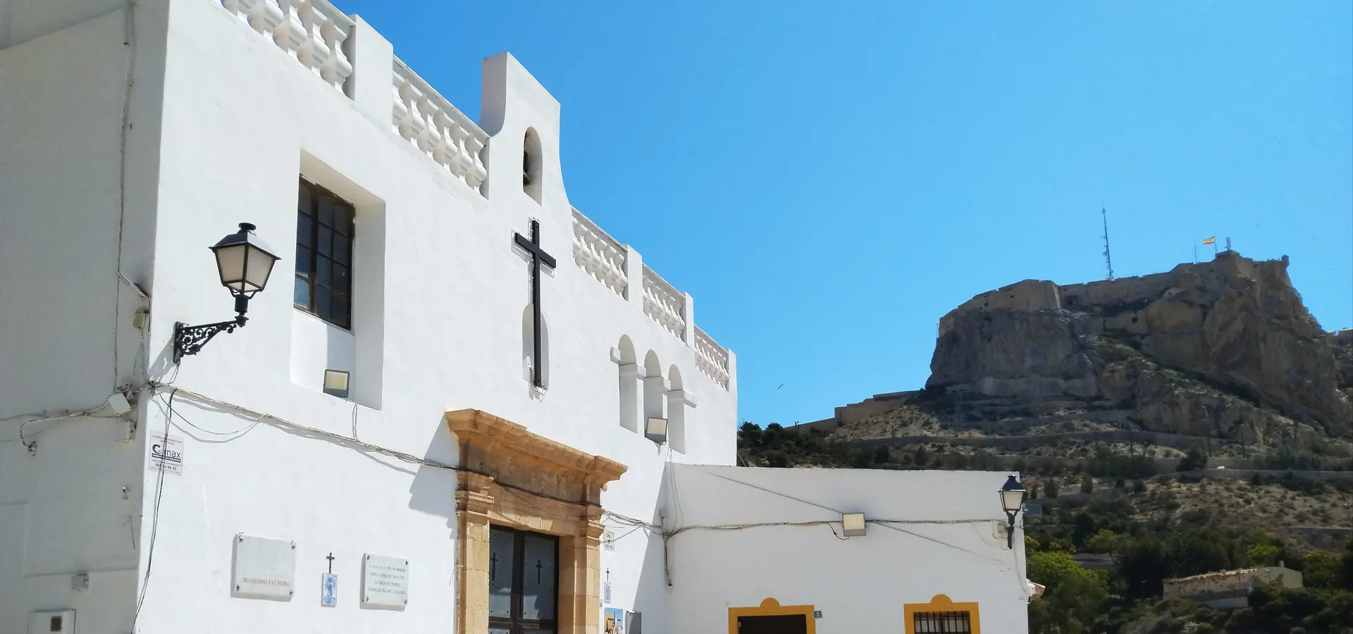 The white Ermita de Santa Cruz, with a black cross on its facade and yellow-painted plinth, and the castle on its hill beyond; a general photograph of the hermitage at the top of the quarter