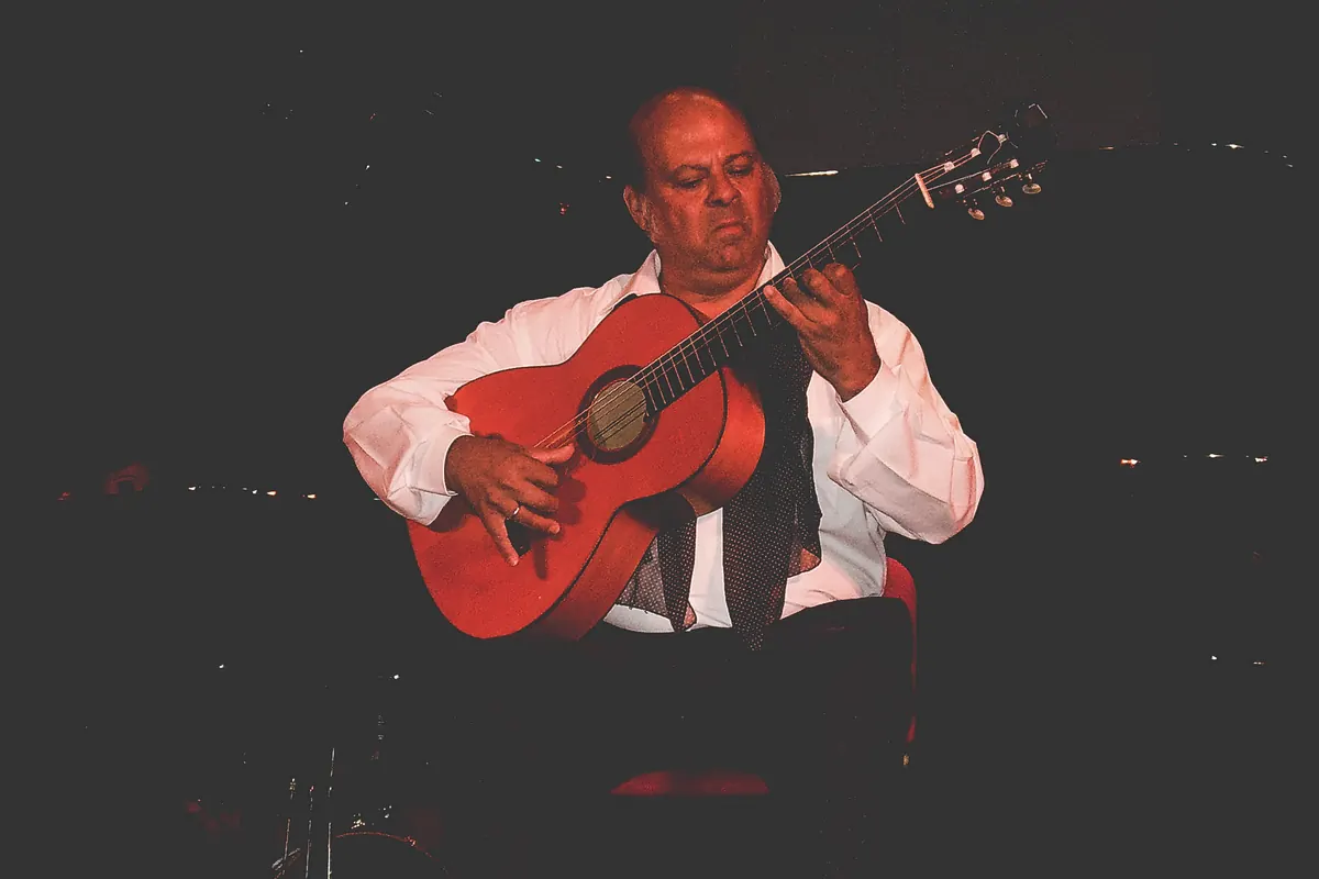 A flamenco guitarist in a white shirt seated on a dark stage, holding a red-toned guitar