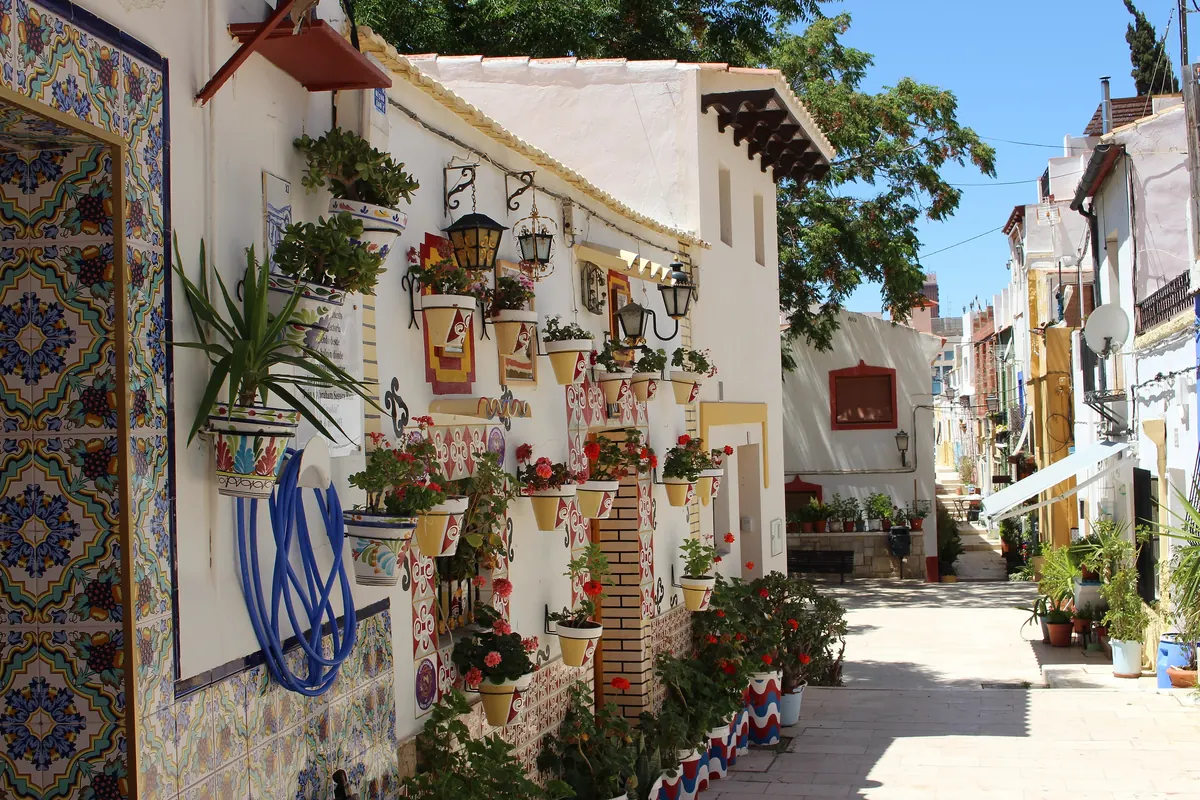 A sunlit lane in Santa Cruz, Alicante, with white walls covered in painted tiles, flower pots and lanterns